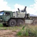 A large military heavy tactical truck, painted in olive drab green, is traveling at speed along a dirt road in an open field environment. The vehicle appears to be a multi-axle logistics or recovery truck, likely a variant of the HEMTT (Heavy Expanded Mobility Tactical Truck) or similar platform, equipped with a crane/loading arm mounted behind the cab. Dust is being kicked up from the rear wheels, and mountains are visible in the background under a partly cloudy sky.