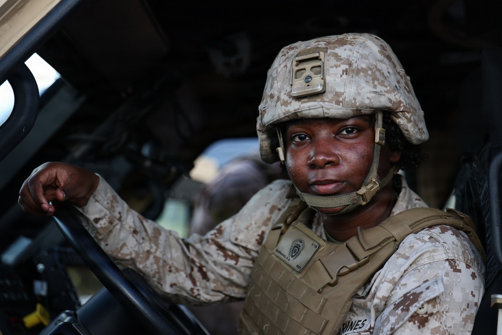 A U.S. Marine in full combat gear is seated in the driver's seat of a military vehicle, looking directly at the camera. The Marine is wearing MARPAT desert camouflage uniform, a combat helmet with a mounted device, and a tan plate carrier vest. The image appears to be a professional military portrait taken in an operational or training environment.