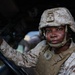 A U.S. Marine in full combat gear is seated in the driver's seat of a military vehicle, looking directly at the camera. The Marine is wearing MARPAT desert camouflage uniform, a combat helmet with a mounted device, and a tan plate carrier vest. The image appears to be a professional military portrait taken in an operational or training environment.