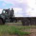 A military flatbed cargo truck in woodland camouflage paint scheme is traveling at speed across an open, semi-arid terrain, kicking up dust. The vehicle appears to be a Medium Tactical Vehicle Replacement (MTVR) or similar U.S. military truck with a flatbed configuration and no cargo loaded. The image is captured with motion blur, suggesting the truck is moving rapidly across what appears to be a training or operational area.