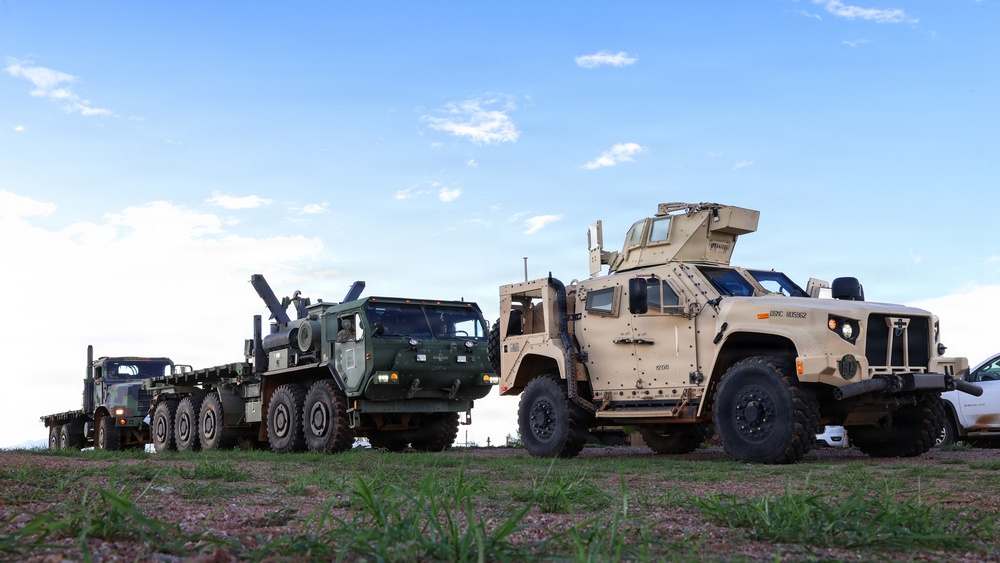 The image shows a lineup of U.S. military vehicles parked on a grassy field under a partly cloudy sky. Visible are a tan-colored Joint Light Tactical Vehicle (JLTV) in the foreground and a green heavy military truck (likely an HEMTT or similar 8-wheeled tactical truck) pulling a flatbed trailer with what appears to be an artillery piece or weapon system loaded on it. The scene appears to be a staging or display area, possibly at a military exercise or base.