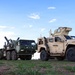 The image shows a lineup of U.S. military vehicles parked on a grassy field under a partly cloudy sky. Visible are a tan-colored Joint Light Tactical Vehicle (JLTV) in the foreground and a green heavy military truck (likely an HEMTT or similar 8-wheeled tactical truck) pulling a flatbed trailer with what appears to be an artillery piece or weapon system loaded on it. The scene appears to be a staging or display area, possibly at a military exercise or base.