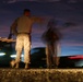 A long-exposure night photograph showing at least two individuals in military camouflage uniforms standing near a roadway at dusk or dawn. The extended shutter speed has created motion blur on one figure and streaking light trails from passing vehicles. The low-angle ground-level perspective and twilight sky create a dramatic compositional effect.