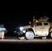A night-time long-exposure photograph of a tan/desert-colored military tactical vehicle, identifiable as a USMC Joint Light Tactical Vehicle (JLTV), parked on a gravel surface. The long exposure has captured motion blur of one or more personnel moving in front of the vehicle on the left side. The vehicle's headlights, amber marker lights, and red tail lights are illuminated, creating light streaks characteristic of long-exposure photography.
