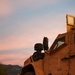 A tan/desert-colored Mine-Resistant Ambush Protected (MRAP) military vehicle is parked on a dirt road during golden hour or sunset. The vehicle bears the identification number M28349 and multiple stenciled warnings including 'NO STEP' and 'NO LIFT'. The background features a mountainous landscape with vegetation under a pastel-toned sky.