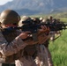 A line of military personnel in desert camouflage uniforms are aiming rifles in a coordinated firing line formation. The soldiers are equipped with tactical gear including helmets, body armor, and optical sights mounted on their weapons. The setting appears to be an outdoor training range with green grass and mountains visible in the background.