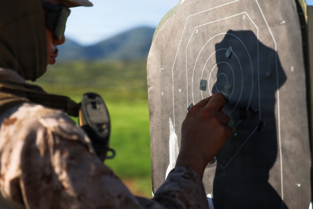 A military personnel wearing desert camouflage uniform and tactical hearing protection is examining or patching a paper shooting target at an outdoor firing range. The target shows multiple bullet holes concentrated in the center and surrounding scoring zones. The background reveals a green, hilly landscape under clear skies.