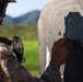 A military personnel wearing desert camouflage uniform and tactical hearing protection is examining or patching a paper shooting target at an outdoor firing range. The target shows multiple bullet holes concentrated in the center and surrounding scoring zones. The background reveals a green, hilly landscape under clear skies.
