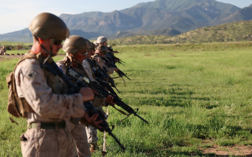 A line of military personnel in desert camouflage uniforms and combat helmets are standing in formation on a grassy field, holding rifles in a ready position. The setting appears to be a training exercise or range qualification event, with mountainous terrain visible in the background. The foreground soldier is in sharp focus while the line recedes into the background.