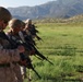 A line of military personnel in desert camouflage uniforms and combat helmets are standing in formation on a grassy field, holding rifles in a ready position. The setting appears to be a training exercise or range qualification event, with mountainous terrain visible in the background. The foreground soldier is in sharp focus while the line recedes into the background.