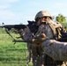 The image shows multiple military personnel in a firing line formation, aiming rifles in the same direction across an open grassy field. The soldiers are wearing desert MARPAT camouflage uniforms and tactical gear consistent with U.S. Marine Corps equipment. The scene appears to be a training exercise or range qualification event.