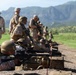 Military personnel in desert camouflage uniforms are conducting a rifle range training exercise. Several soldiers are in the prone position firing rifles downrange while others stand behind them, likely acting as instructors or awaiting their turn. The setting appears to be an outdoor firing range with mountainous terrain visible in the background.