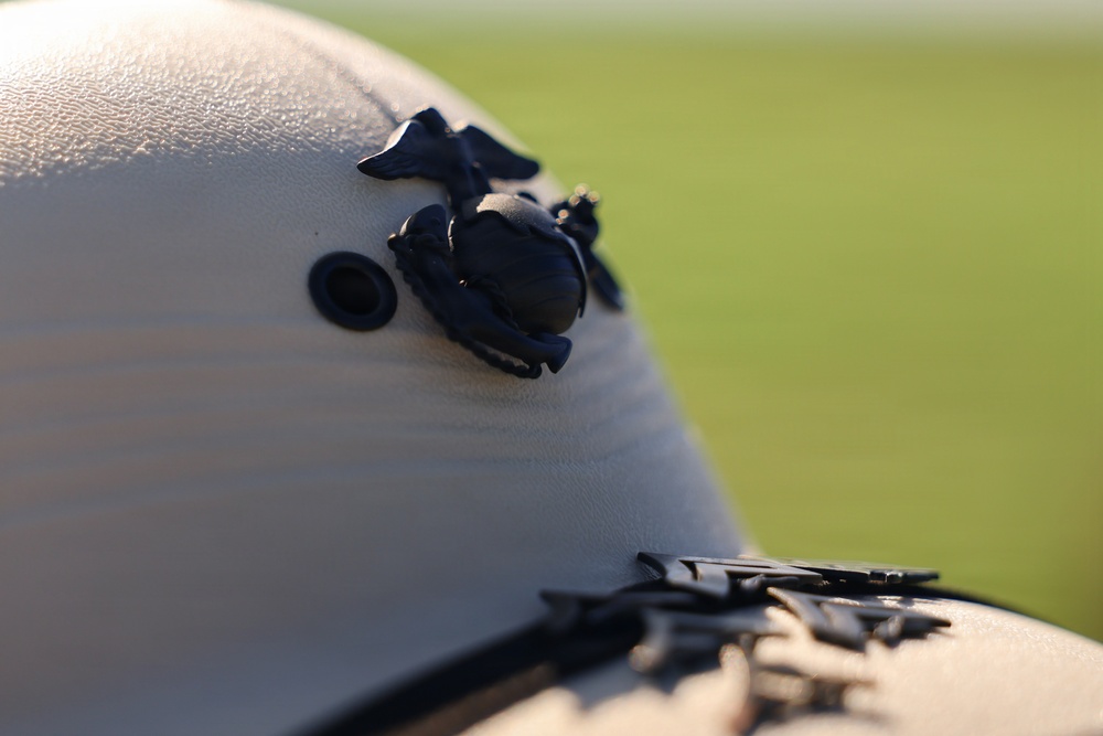 A close-up photograph of a white military-style helmet featuring a dark metal Eagle, Globe, and Anchor (EGA) emblem, which is the insignia of the United States Marine Corps. The helmet also has a visible grommet hole and a chin strap buckle at the bottom edge. The background is blurred, showing what appears to be a green grassy field in soft focus.