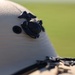 A close-up photograph of a white military-style helmet featuring a dark metal Eagle, Globe, and Anchor (EGA) emblem, which is the insignia of the United States Marine Corps. The helmet also has a visible grommet hole and a chin strap buckle at the bottom edge. The background is blurred, showing what appears to be a green grassy field in soft focus.
