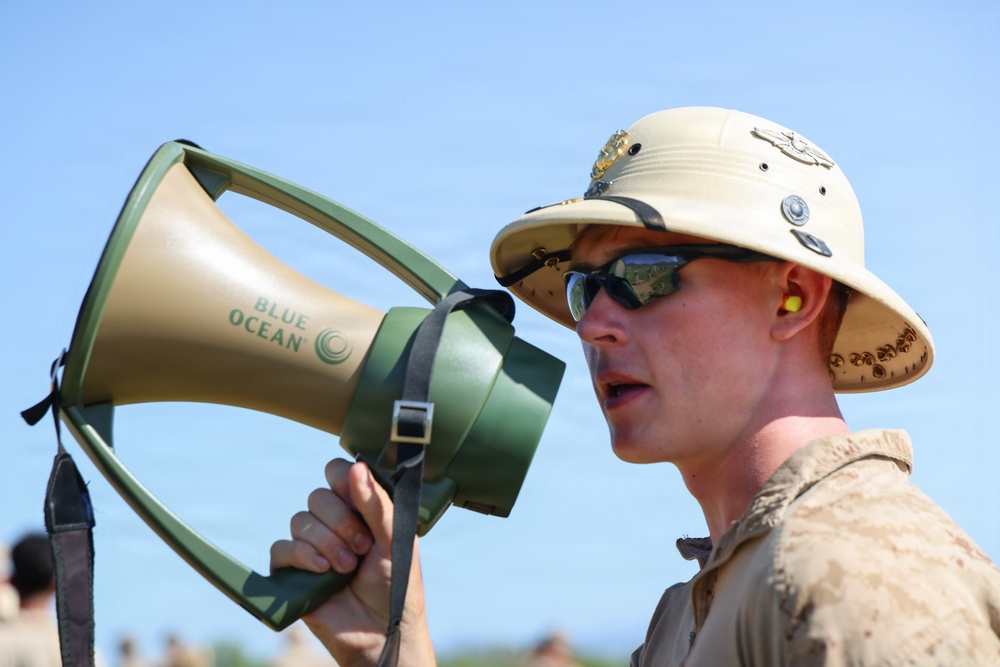 A military personnel member is speaking into a 'Blue Ocean' brand megaphone/bullhorn in an outdoor setting. The individual is wearing a tan pith helmet with insignia badges, reflective sunglasses, yellow ear protection, and a tan military uniform. The image is a close-up shot taken under clear blue sky conditions.