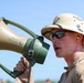 A military personnel member is speaking into a 'Blue Ocean' brand megaphone/bullhorn in an outdoor setting. The individual is wearing a tan pith helmet with insignia badges, reflective sunglasses, yellow ear protection, and a tan military uniform. The image is a close-up shot taken under clear blue sky conditions.