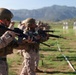 A group of military personnel in desert camouflage uniforms are conducting a rifle marksmanship training exercise on an outdoor firing range. Multiple shooters are lined up in a row, aiming rifles downrange toward white target stands. The setting appears to be a military training facility with mountainous terrain visible in the background.
