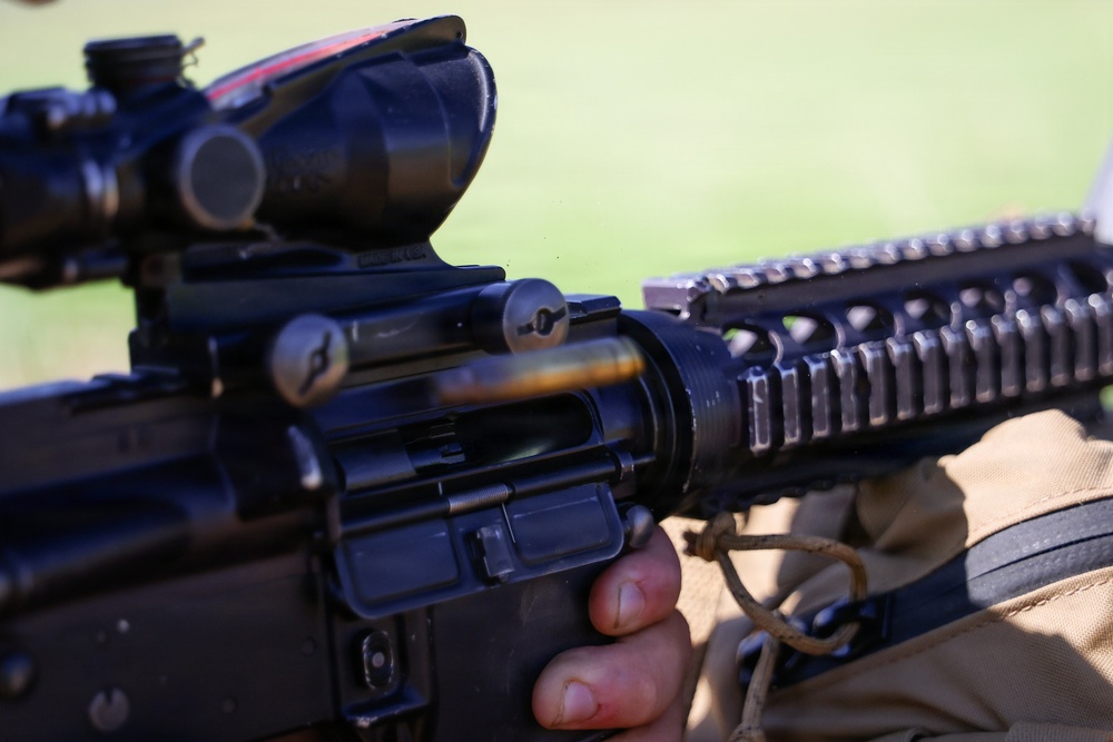 A close-up photograph of a military-style rifle being held by a person wearing tan/khaki tactical clothing. The weapon is equipped with a magnified optic scope (consistent with a ACOG-style sight) and a Picatinny rail handguard system. The image is taken outdoors with a green background, likely a field or range environment.