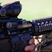 A close-up photograph of a military-style rifle being held by a person wearing tan/khaki tactical clothing. The weapon is equipped with a magnified optic scope (consistent with a ACOG-style sight) and a Picatinny rail handguard system. The image is taken outdoors with a green background, likely a field or range environment.
