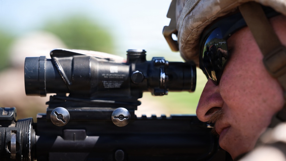 A military service member is shown in close-up profile view, aiming down the optic sight of a rifle. The individual is wearing a tan/desert camouflage helmet and tactical sunglasses, with a reflection visible in the lens. The image is a sharp, high-resolution photograph focused on the weapon system and shooter's face.