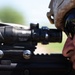 A military service member is shown in close-up profile view, aiming down the optic sight of a rifle. The individual is wearing a tan/desert camouflage helmet and tactical sunglasses, with a reflection visible in the lens. The image is a sharp, high-resolution photograph focused on the weapon system and shooter's face.