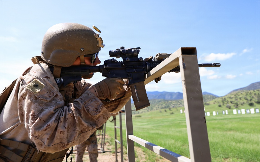 A U.S. Marine in full combat gear is firing an M4 carbine rifle at a military shooting range. A spent brass casing is visibly ejecting from the weapon's ejection port. The range features multiple target stands extending into the distance against a backdrop of green hills and blue sky.