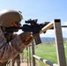 A U.S. Marine in full combat gear is firing an M4 carbine rifle at a military shooting range. A spent brass casing is visibly ejecting from the weapon's ejection port. The range features multiple target stands extending into the distance against a backdrop of green hills and blue sky.