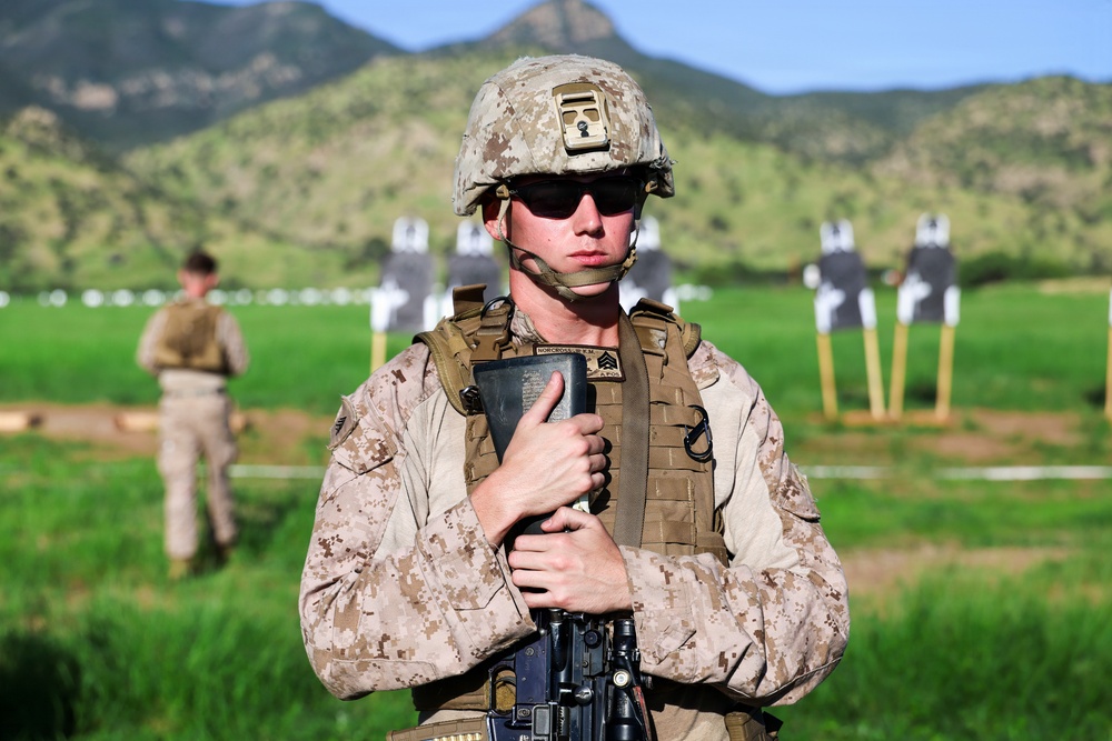 A U.S. Marine in desert MARPAT camouflage uniform stands at what appears to be an outdoor rifle range, holding a notebook or clipboard against their chest. The Marine is wearing a combat helmet, tactical vest, sunglasses, and an earpiece, with a rifle visible at the lower frame. Shooting targets and a second service member are visible in the background against a mountainous, green landscape.