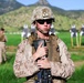 A U.S. Marine in desert MARPAT camouflage uniform stands at what appears to be an outdoor rifle range, holding a notebook or clipboard against their chest. The Marine is wearing a combat helmet, tactical vest, sunglasses, and an earpiece, with a rifle visible at the lower frame. Shooting targets and a second service member are visible in the background against a mountainous, green landscape.