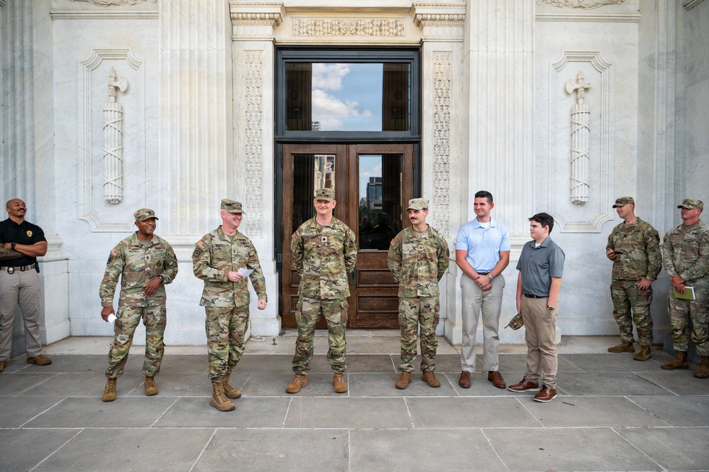 A group of military personnel in OCP (Operational Camouflage Pattern) uniforms and two civilians in casual attire are standing in front of a large ornate neoclassical building with marble columns and decorative pilasters. The individuals appear to be posed for a formal or semi-formal group photo, possibly during an official visit or ceremony. A security officer in a black uniform is also visible on the far left.