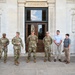A group of military personnel in OCP (Operational Camouflage Pattern) uniforms and two civilians in casual attire are standing in front of a large ornate neoclassical building with marble columns and decorative pilasters. The individuals appear to be posed for a formal or semi-formal group photo, possibly during an official visit or ceremony. A security officer in a black uniform is also visible on the far left.