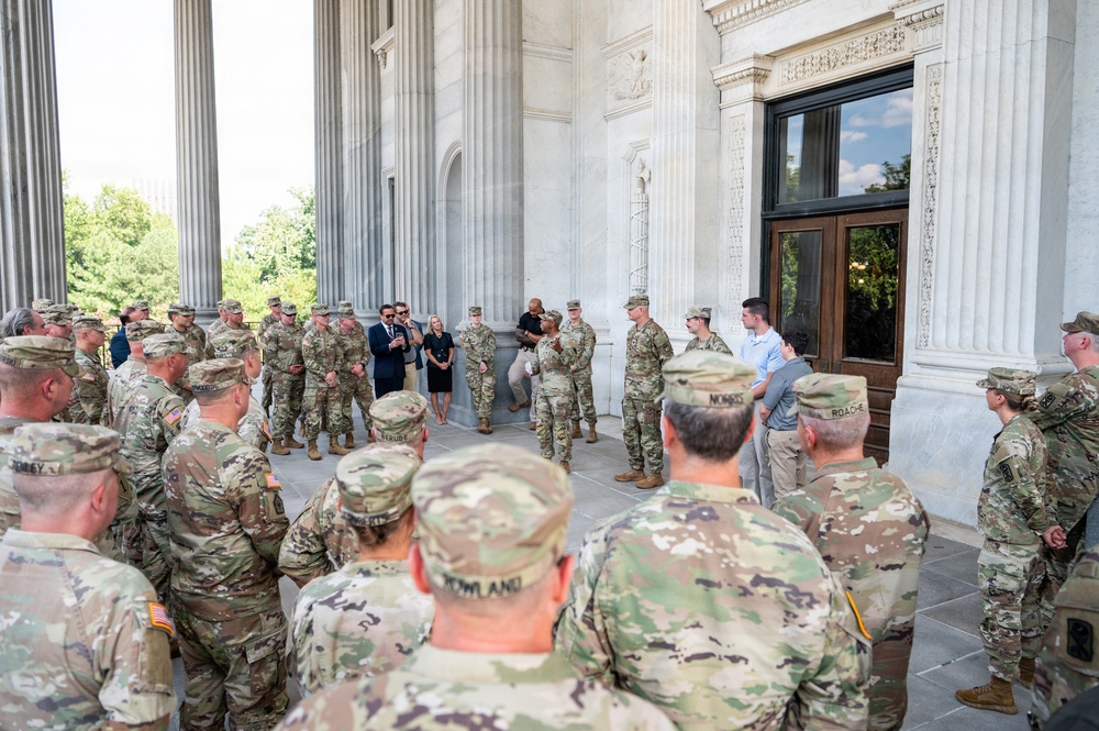 A large group of U.S. Army soldiers in OCP (Operational Camouflage Pattern) uniforms are gathered in a semicircle on the portico of a neoclassical government building. A civilian man in a dark suit appears to be addressing the group, accompanied by a woman in civilian attire. The setting suggests an official briefing, ceremony, or visit at a prominent federal or institutional building.