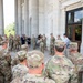 A large group of U.S. Army soldiers in OCP (Operational Camouflage Pattern) uniforms are gathered in a semicircle on the portico of a neoclassical government building. A civilian man in a dark suit appears to be addressing the group, accompanied by a woman in civilian attire. The setting suggests an official briefing, ceremony, or visit at a prominent federal or institutional building.