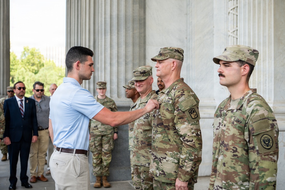 A civilian in a light blue polo shirt is pinning a medal or insignia onto a U.S. Army soldier in multicam/OCP uniform during what appears to be a formal recognition ceremony. The event is taking place in front of large stone columns, consistent with a prominent government or memorial building in Washington D.C. Several other military personnel and civilians in suits are present in the background observing the ceremony.