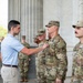 A civilian in a light blue polo shirt is pinning a medal or insignia onto a U.S. Army soldier in multicam/OCP uniform during what appears to be a formal recognition ceremony. The event is taking place in front of large stone columns, consistent with a prominent government or memorial building in Washington D.C. Several other military personnel and civilians in suits are present in the background observing the ceremony.