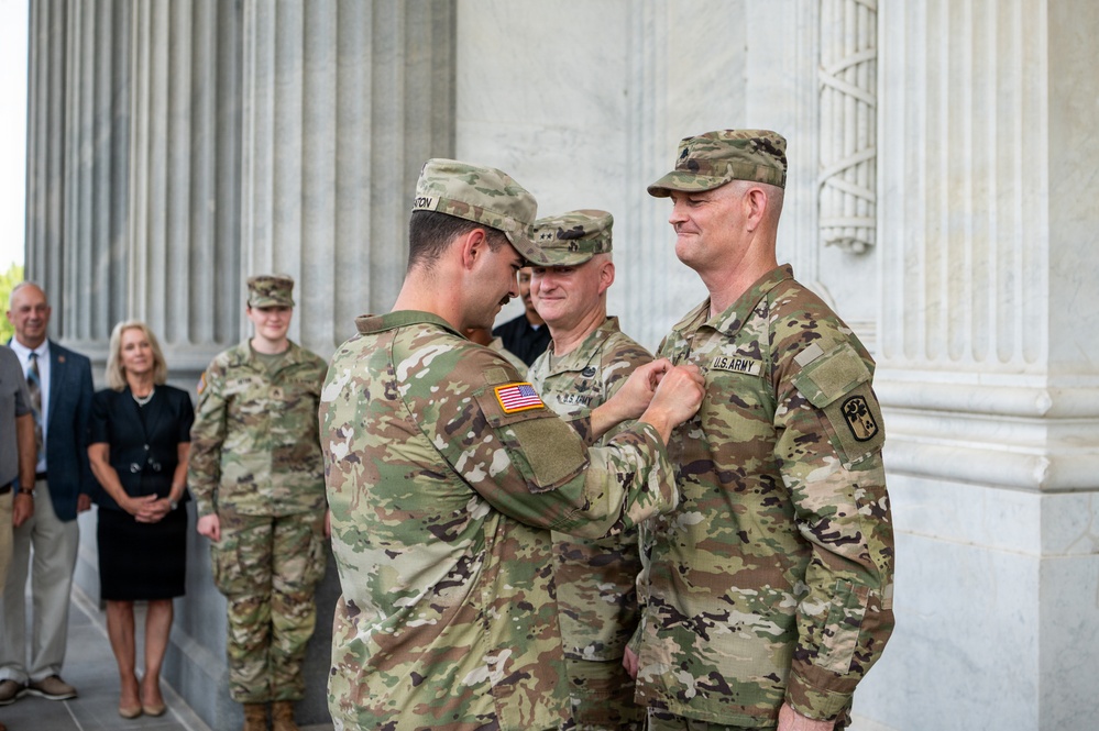 A U.S. Army military ceremony is taking place in front of a large marble columned building. One soldier is pinning a medal or insignia onto the chest of another soldier, while a third uniformed soldier observes closely. Several civilians and another soldier stand in the background as witnesses to the ceremony.