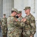 A U.S. Army military ceremony is taking place in front of a large marble columned building. One soldier is pinning a medal or insignia onto the chest of another soldier, while a third uniformed soldier observes closely. Several civilians and another soldier stand in the background as witnesses to the ceremony.