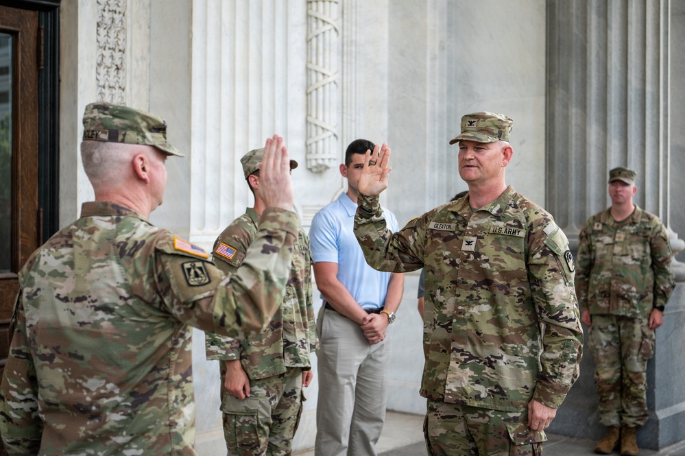 A U.S. Army military ceremony appears to be taking place in front of a neoclassical building with large white columns. Two soldiers in the foreground are raising their right hands, consistent with an oath-taking or promotion ceremony. The soldier on the right is identified by his name tape as 'GLEATON' and wears a U.S. Army colonel's eagle insignia on his cap.