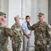 A U.S. Army military ceremony appears to be taking place in front of a neoclassical building with large white columns. Two soldiers in the foreground are raising their right hands, consistent with an oath-taking or promotion ceremony. The soldier on the right is identified by his name tape as 'GLEATON' and wears a U.S. Army colonel's eagle insignia on his cap.