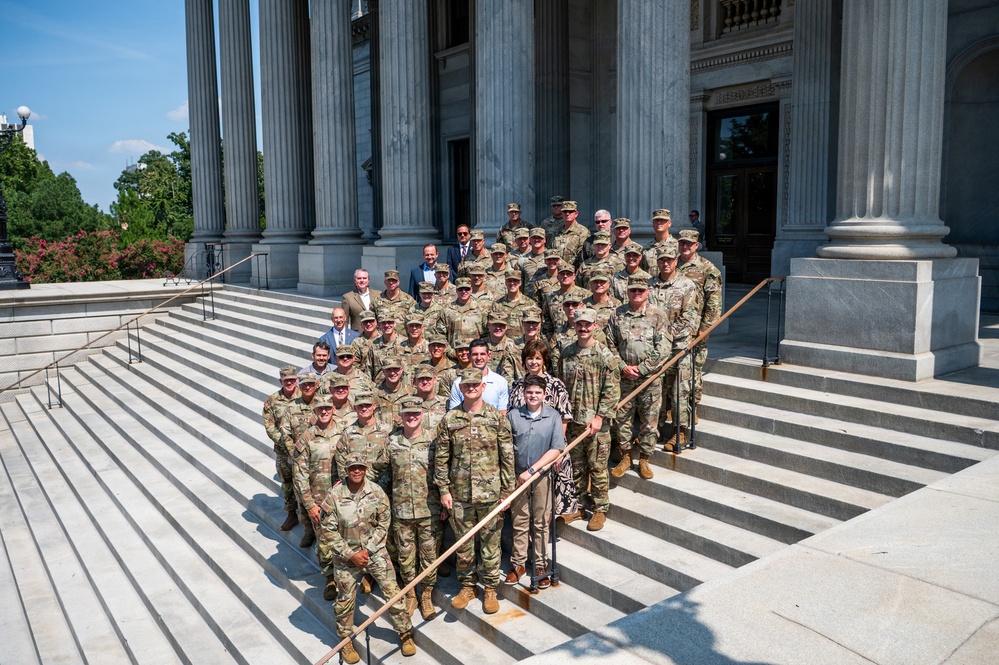 A large group of military personnel in camouflage uniforms, along with several civilians in business and casual attire, pose for a formal group photograph on the steps of a neoclassical government building with large stone columns. The group consists of approximately 35-40 individuals arranged in multiple rows on the wide stone staircase. The setting and attire suggest an official visit or ceremonial occasion at a state capitol or federal building.