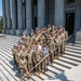 A large group of military personnel in camouflage uniforms, along with several civilians in business and casual attire, pose for a formal group photograph on the steps of a neoclassical government building with large stone columns. The group consists of approximately 35-40 individuals arranged in multiple rows on the wide stone staircase. The setting and attire suggest an official visit or ceremonial occasion at a state capitol or federal building.