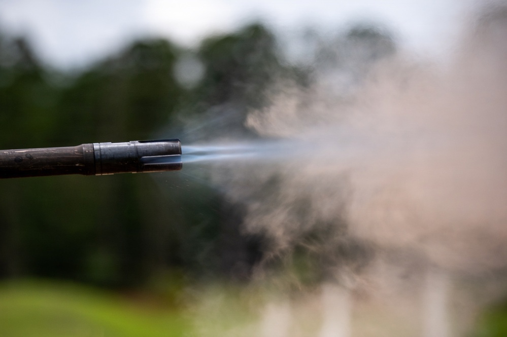 The image captures a close-up of a firearm muzzle at the moment of discharge, showing propellant gases and smoke expelled from the barrel. A flash hider or muzzle device is visible at the end of the barrel. The background is blurred with green foliage and an overcast sky.
