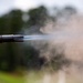 The image captures a close-up of a firearm muzzle at the moment of discharge, showing propellant gases and smoke expelled from the barrel. A flash hider or muzzle device is visible at the end of the barrel. The background is blurred with green foliage and an overcast sky.