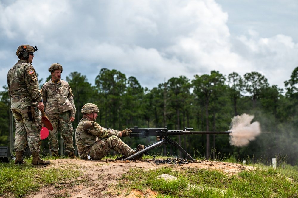 Three military personnel in camouflage uniforms are conducting a live-fire training exercise at an outdoor range. One soldier is in a seated firing position operating what appears to be an M2 .50 caliber heavy machine gun mounted on a tripod, with visible muzzle blast and smoke. Two other soldiers stand nearby observing, one holding a red range paddle.