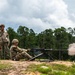 Three military personnel in camouflage uniforms are conducting a live-fire training exercise at an outdoor range. One soldier is in a seated firing position operating what appears to be an M2 .50 caliber heavy machine gun mounted on a tripod, with visible muzzle blast and smoke. Two other soldiers stand nearby observing, one holding a red range paddle.