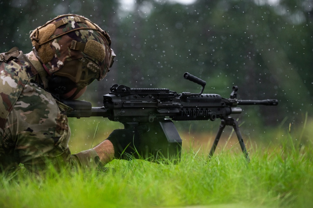 A soldier in multicam camouflage uniform is in a prone firing position in a grassy field during rainfall. The soldier is aiming what appears to be an M249 Squad Automatic Weapon (SAW) or similar light machine gun mounted on a bipod. The image captures a training or operational scenario in wet, outdoor conditions.