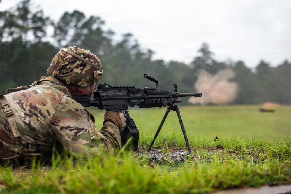 A soldier in MultiCam/OCP camouflage uniform is firing a light machine gun from a prone position on a grass field. The weapon is mounted on a bipod and appears to be actively firing, as evidenced by a dust/debris cloud visible downrange. The setting appears to be a military training range during overcast, possibly rainy conditions.