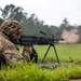 A soldier in MultiCam/OCP camouflage uniform is firing a light machine gun from a prone position on a grass field. The weapon is mounted on a bipod and appears to be actively firing, as evidenced by a dust/debris cloud visible downrange. The setting appears to be a military training range during overcast, possibly rainy conditions.