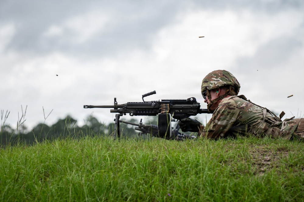 A soldier in full combat gear is in a prone firing position on a grassy berm, actively firing a light machine gun. Multiple brass shell casings are visibly ejecting through the air, indicating live fire. A second weapon system is partially visible in the background, suggesting a range training exercise.
