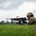 A soldier in full combat gear is in a prone firing position on a grassy berm, actively firing a light machine gun. Multiple brass shell casings are visibly ejecting through the air, indicating live fire. A second weapon system is partially visible in the background, suggesting a range training exercise.